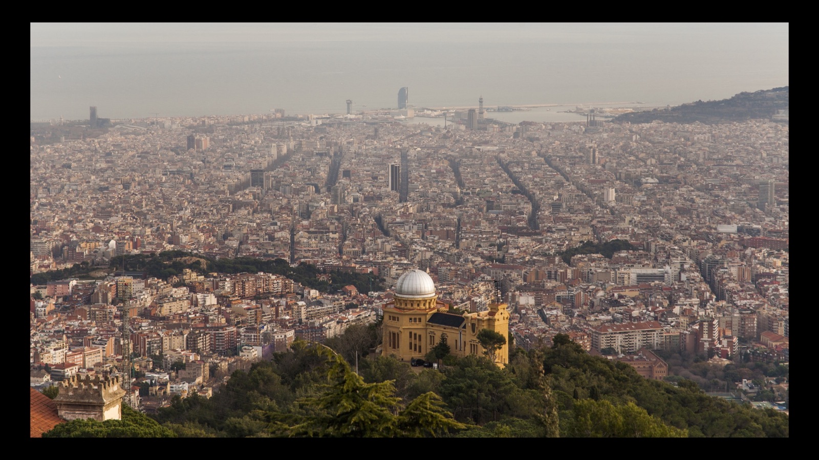 La cúpula platejada de l'Observatori Fabra sobre Barcelona, amb la trama de la ciutat estenent-se fins al port i el mar dissolent-se en la calitja