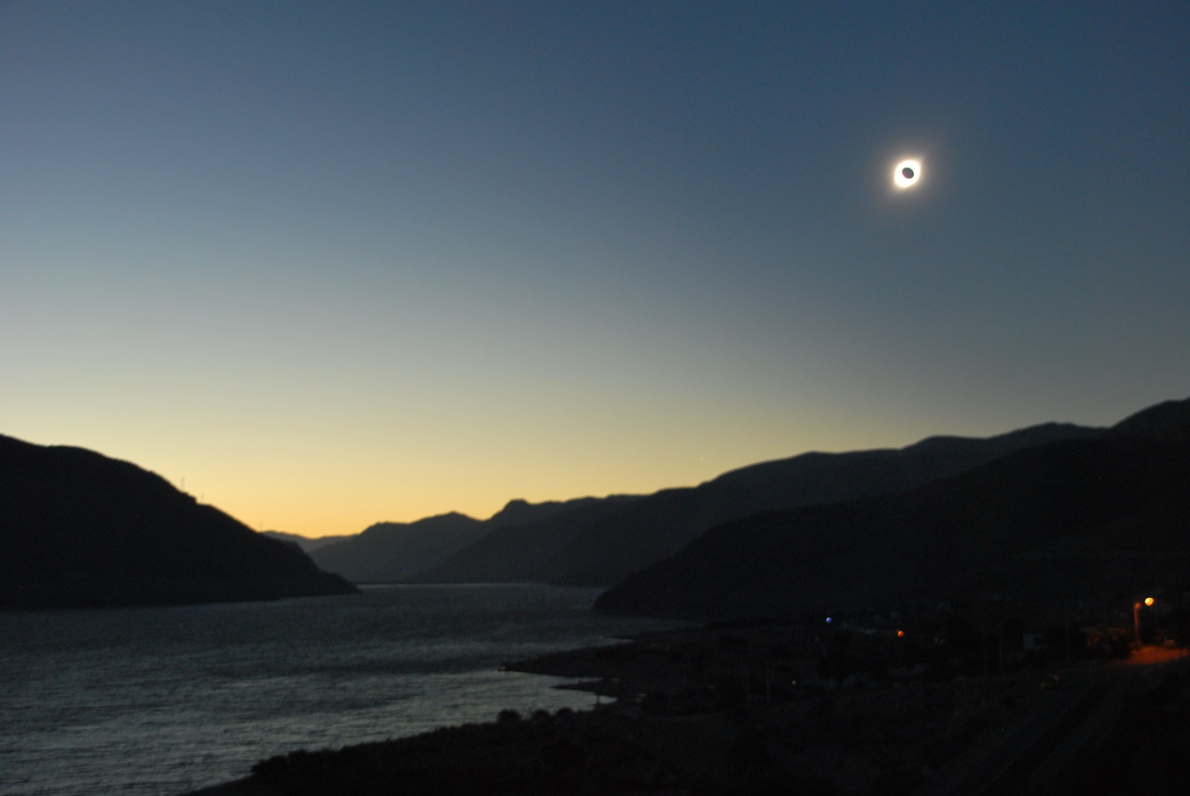 El Sol totalment eclipsat, amb la corona i l'anell de diamant, baix sobre un embassament envoltat de muntanyes al crepuscle a Xile, 2019